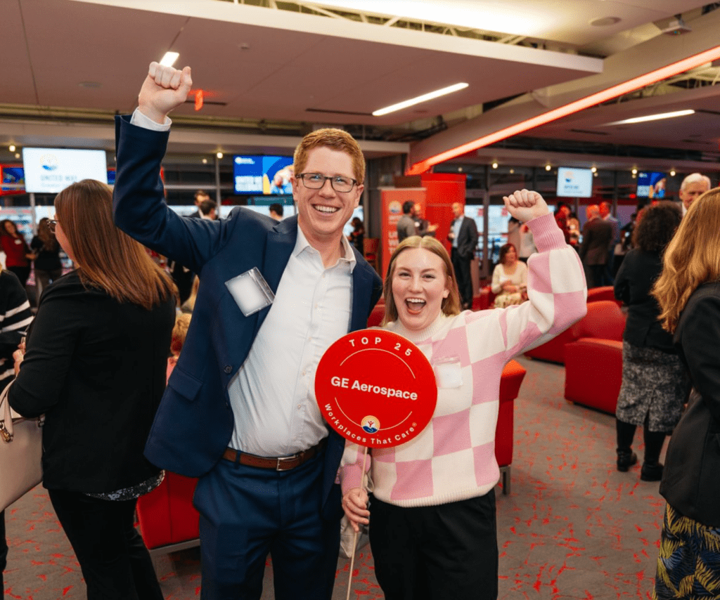 Two people at an indoor event raise their arms and smile while holding a red sign that reads “Top 25 GE Aerospace Workplaces That Care,” with attendees and seating visible in the background.