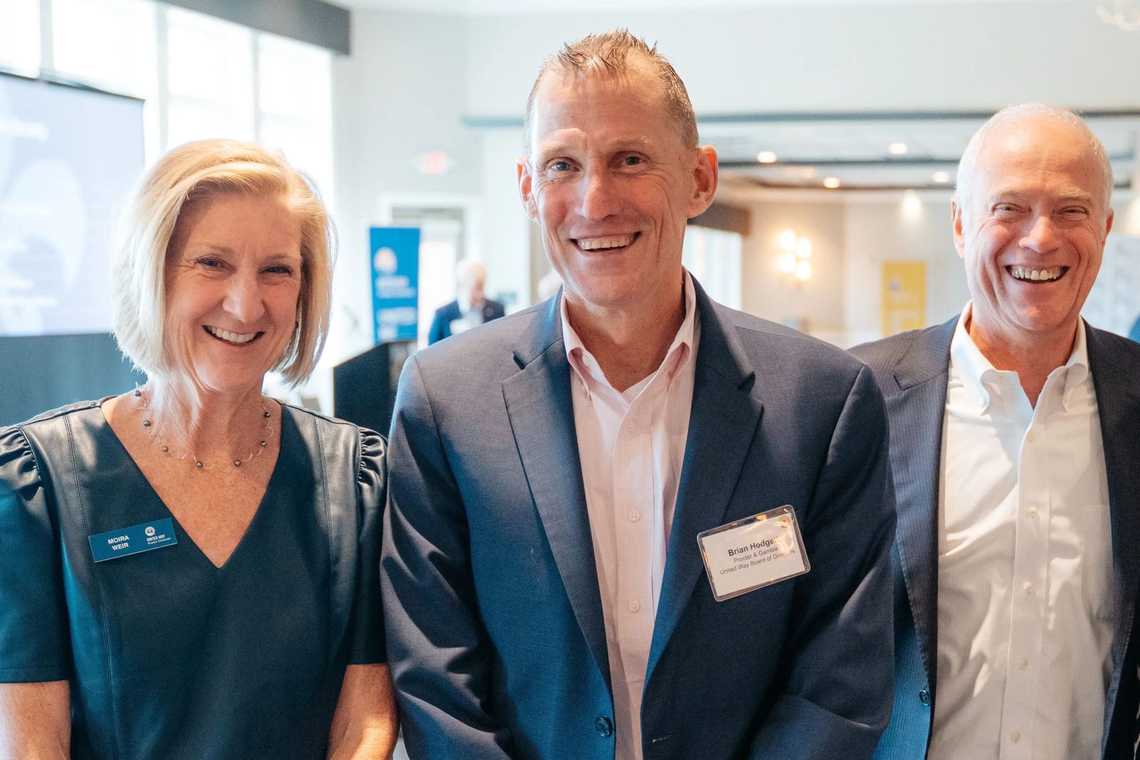 Three people in business attire wearing name badges smile at an indoor networking event.