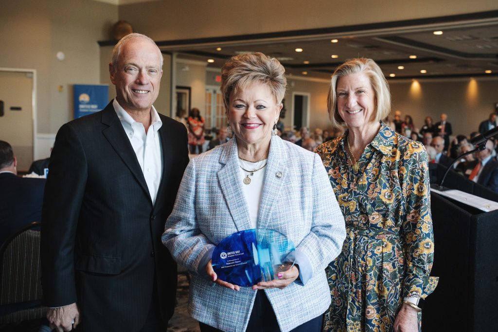 Three adults smile at an indoor event, with the woman in the center holding a blue glass award.