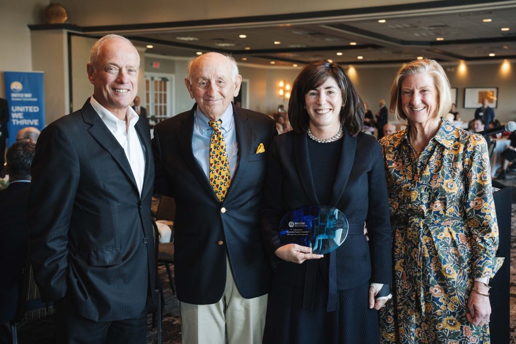Four adults smile at an indoor event, with a woman holding a blue glass award.