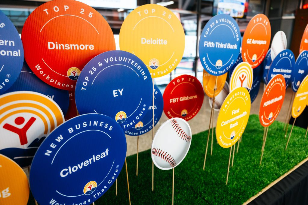 Colorful circular signs on sticks displayed in artificial grass with company names such as Dinsmore, Deloitte, EY, and Fifth Third Bank under “Top 25” workplace categories, with a baseball prop in front.