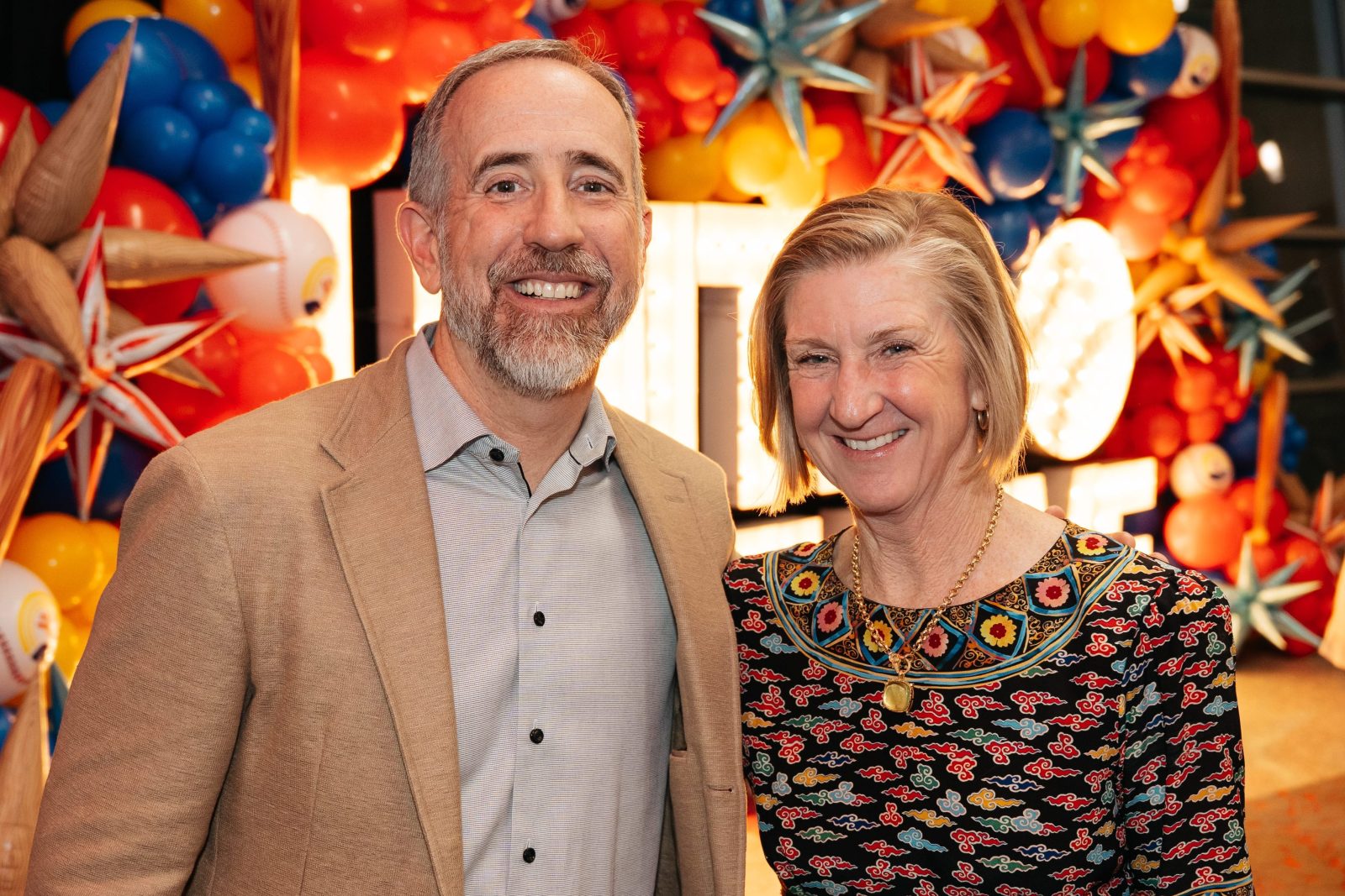 Two people smiling and posing in front of a colorful balloon and star decoration display at an indoor event.