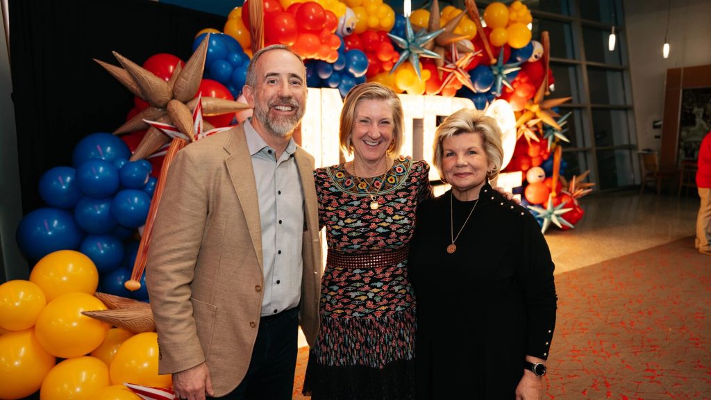 Three people smiling and posing in front of a colorful balloon and star decoration display at an indoor event.