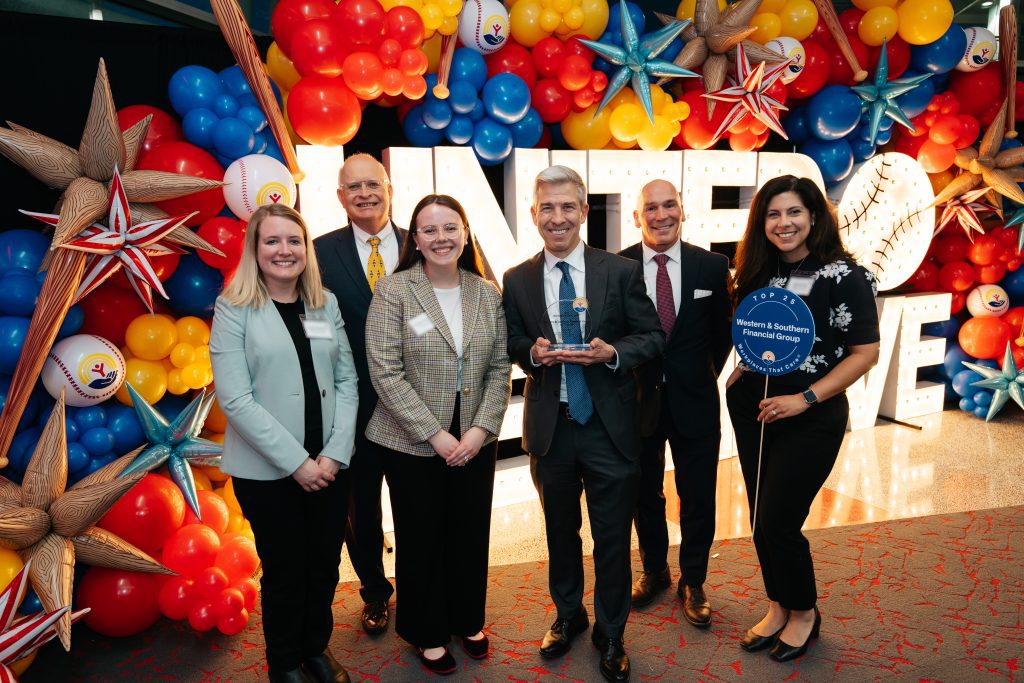 Six people posing in front of a colorful balloon display and large “UNITED WE THRIVE” letters, with one person holding a “Top 25 Western & Southern Financial Group Workplaces That Care” sign and another holding an award.