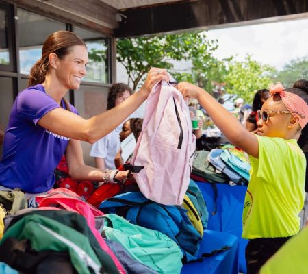 Volunteers distribute colorful backpacks at an outdoor community event, with one person in a purple shirt handing a pink backpack to a child in a yellow shirt, surrounded by tables and trees.